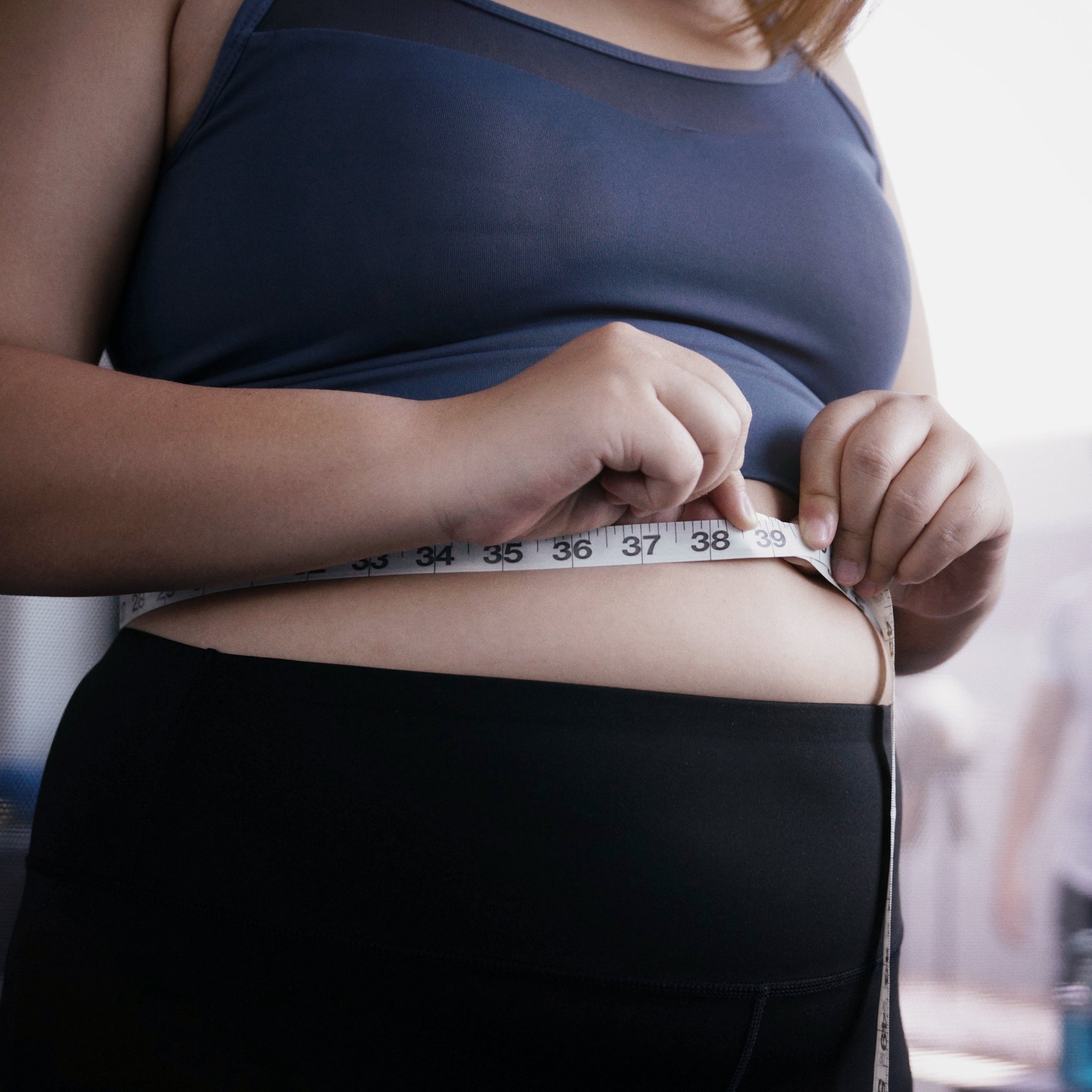 A person in a dark sports bra and black leggings measures their waist with a tape measure, focusing on the midsection
