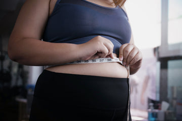 A person in a dark sports bra and black leggings measures their waist with a tape measure, focusing on the midsection
