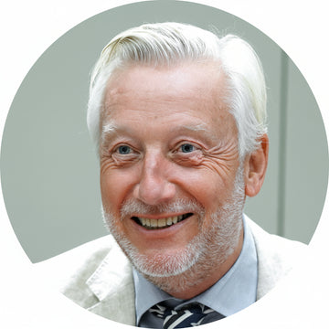 Man with white hair and beard wearing a light-colored suit against a neutral background